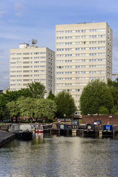 Típico edifício alto bloco de apartamentos, localizado em uma cidade inglesa. O edifício está localizado com vista para um canal, que tem barcos de canal ancorados na margem. O tempo está ensolarado
 - Foto, Imagem
