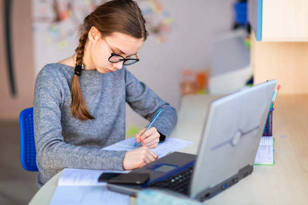 Beautiful young school girl left-handed working at home in her room with a laptop and class notes studying in a virtual class. Distance education and learning, e-learning, online learning concept during quarantine - Φωτογραφία, εικόνα