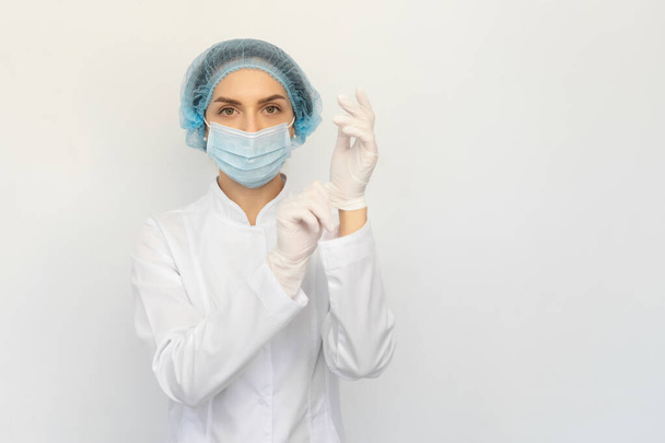 Женщина-врач в медицинской маске надевает белые резиновые перчатки
 - Фото, изображение