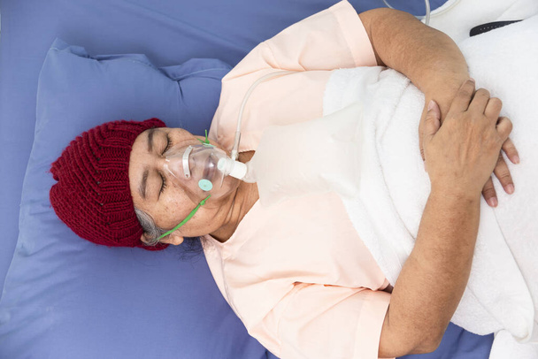 Asiatische ältere Frau trägt Wollmütze mit Sauerstoffmaske zum Atmen, während sie auf dem Patientenbett liegt, Seniormedizin- und Gesundheitskonzept - Foto, Bild