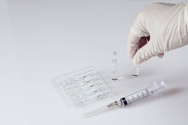 Disposable syringe, upright ampoules with medicine and ampoules with medicine in a blister on white acrylic table, hand in white medical glove picking up one ampoule - Valokuva, kuva