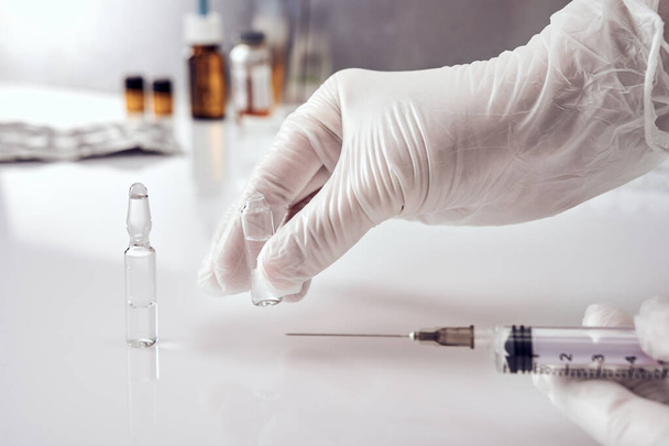 Female hands in white medical gloves, right hand picking up one of two ampoules with medicine from white acrylic table, left hand holding a syringe. Ampoules, glass jars, blisters on background - 写真・画像