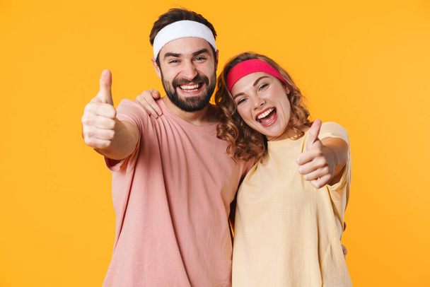 Portrait of athletic young couple wearing headbands smiling and gesturing thumbs up isolated over yellow background - Foto, afbeelding
