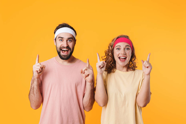 Portrait of athletic young couple wearing headbands smiling and pointing fingers at copyspace isolated over yellow background - Фото, изображение