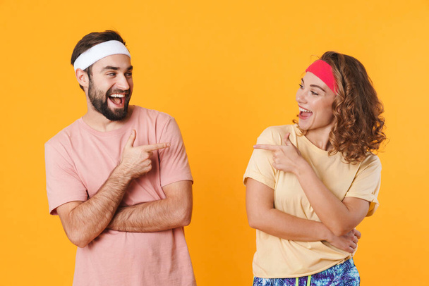 Portrait of athletic young couple wearing headbands smiling and pointing fingers at each other isolated over yellow background - Foto, Imagen