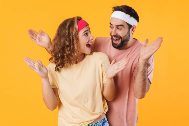 Portrait of athletic young couple wearing headbands smiling to each other and expressing surprise isolated over yellow background - Photo, Image