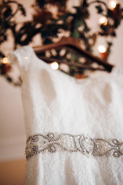 Біла мереживна весільна сукня з поясом, вишитим камінням, висить на дизайнерській люстрі
 - Фото, зображення