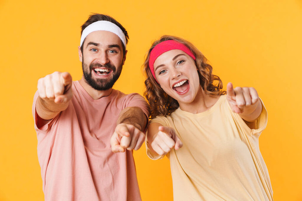 Portrait of athletic young couple wearing headbands smiling and pointing fingers at camera isolated over yellow background - Foto, immagini