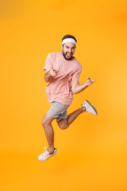 Image of muscular athletic young man in shorts and t-shirt having fun while doing workout isolated over yellow background - 写真・画像