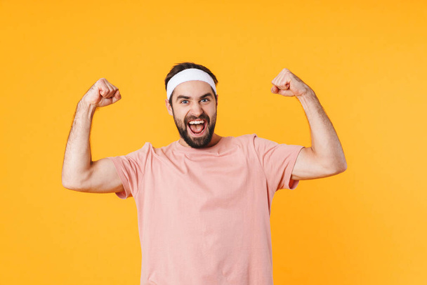 Image of muscular athletic young man in t-shirt having fun and showing his biceps isolated over yellow background - 写真・画像