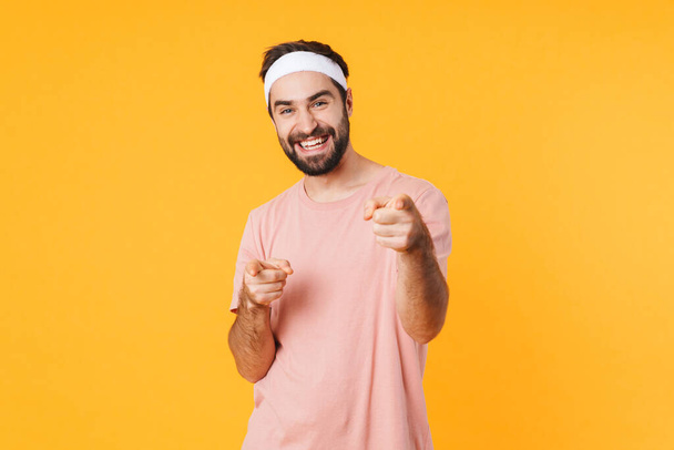 Image of muscular athletic young man in t-shirt smiling and pointing fingers isolated over yellow background - Foto, Bild