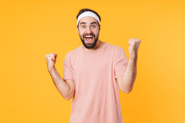 Image of muscular athletic young man in t-shirt smiling and clenching fists in victory isolated over yellow background - Foto, imagen