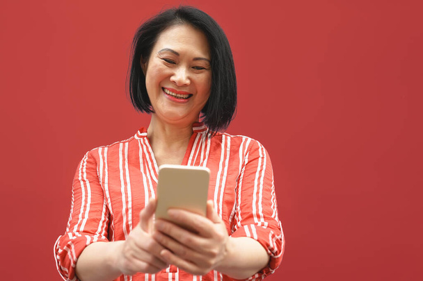 Ευτυχισμένη Ασιάτισσα γυναίκα που χρησιμοποιεί κινητό smartphone υπαίθρια - Γυναίκα ανώτερος διασκεδάζει με τις νέες μοντέρνες εφαρμογές κοινωνικής δικτύωσης - Ηλικιωμένοι τρόπο ζωής και τεχνολογίας ηλικιωμένοι εθισμένοι έννοια - Κόκκινο φόντο - Φωτογραφία, εικόνα