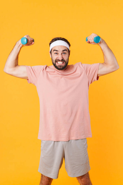 Image of muscular athletic young man in t-shirt having fun and lifting dumbbells isolated over yellow background - Фото, изображение