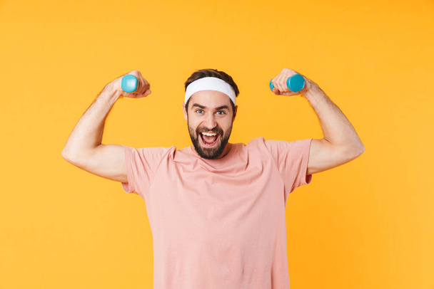 Image of muscular athletic young man in t-shirt having fun and lifting dumbbells isolated over yellow background - 写真・画像