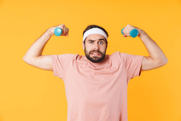 Image of muscular athletic young man in t-shirt having fun and lifting dumbbells isolated over yellow background - Fotó, kép