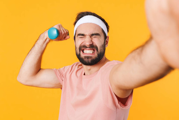 Image of muscular athletic young man in t-shirt taking selfie photo while lifting dumbbell isolated over yellow background - Photo, image