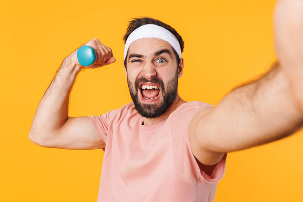 Image of muscular athletic young man in t-shirt taking selfie photo while lifting dumbbell isolated over yellow background - Φωτογραφία, εικόνα
