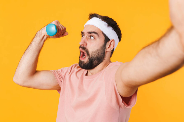 Image of muscular athletic young man in t-shirt taking selfie photo while lifting dumbbell isolated over yellow background - Valokuva, kuva