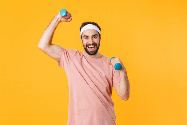 Image of muscular athletic young man in t-shirt having fun and lifting dumbbells isolated over yellow background - Photo, Image
