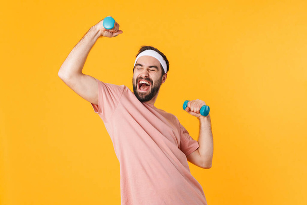 Image of muscular athletic young man in t-shirt having fun and lifting dumbbells isolated over yellow background - Fotoğraf, Görsel