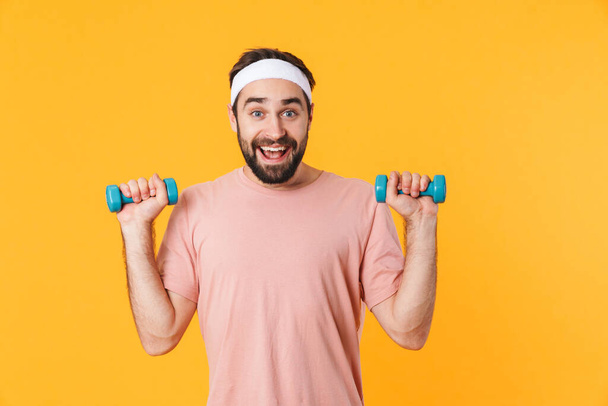Image of muscular athletic young man in t-shirt having fun and lifting dumbbells isolated over yellow background - Photo, image
