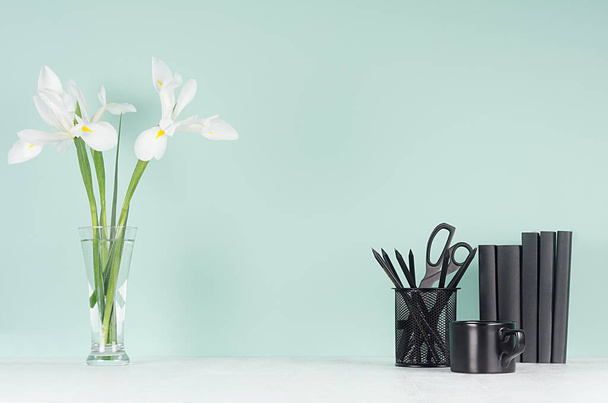 Minimalistic style of home workplace with black stationery, books, coffee cup, fresh white spring flowers on green mint menthe wall and white wood board. - Fotó, kép