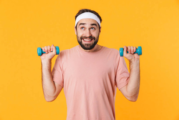 Image of muscular athletic young man in t-shirt having fun and lifting dumbbells isolated over yellow background - Foto, afbeelding