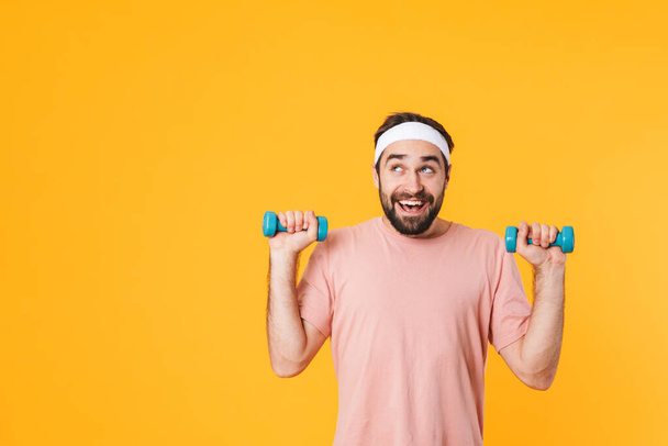 Image of muscular athletic young man in t-shirt having fun and lifting dumbbells isolated over yellow background - Fotografie, Obrázek