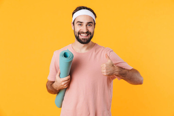 Image of muscular athletic young man in t-shirt smiling and carrying fitness mat isolated over yellow background - Foto, imagen