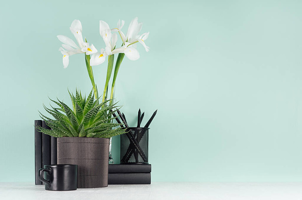 Cozy home workplace with black stationery, books, coffee cup, green aloe, fresh spring white iris bouquet in trend green mint menthe color interior and white wood table. - Fotó, kép