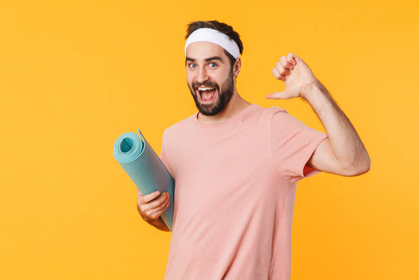 Image of muscular athletic young man in t-shirt smiling and carrying fitness mat isolated over yellow background - Foto, afbeelding