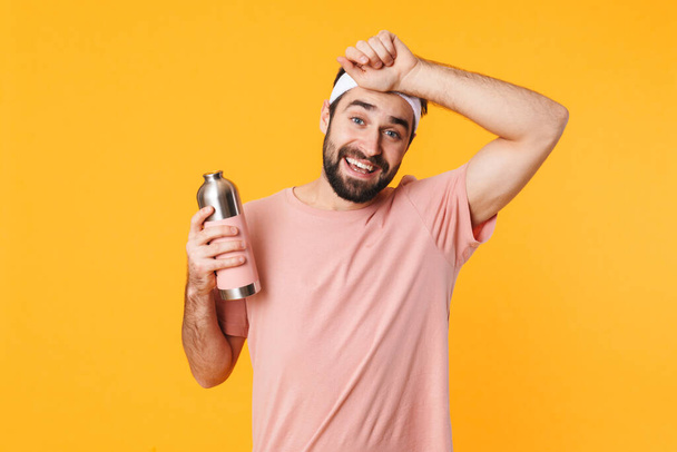 Image of muscular athletic young man in t-shirt holding water bottle isolated over yellow background - Fotoğraf, Görsel
