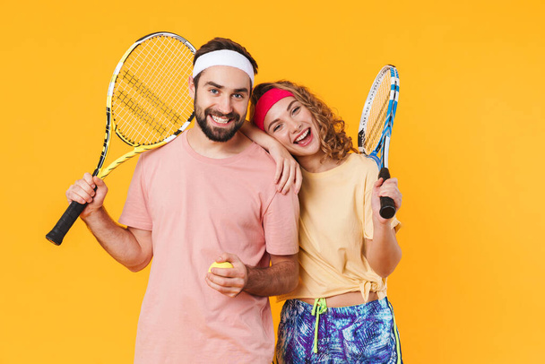 Portrait of athletic young happy couple wearing headbands holding rackets while playing tennis together isolated over yellow background - Photo, Image