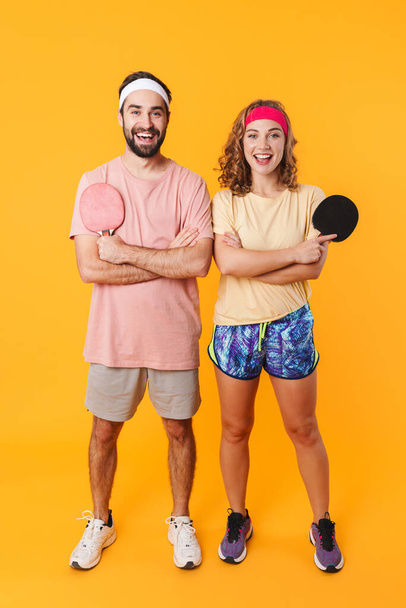 Portrait of athletic young couple wearing headbands holding rackets while playing table tennis together isolated over yellow background - Fotó, kép