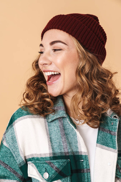 Photo of pretty joyful woman in knit hat laughing and looking aside isolated over beige background - Zdjęcie, obraz