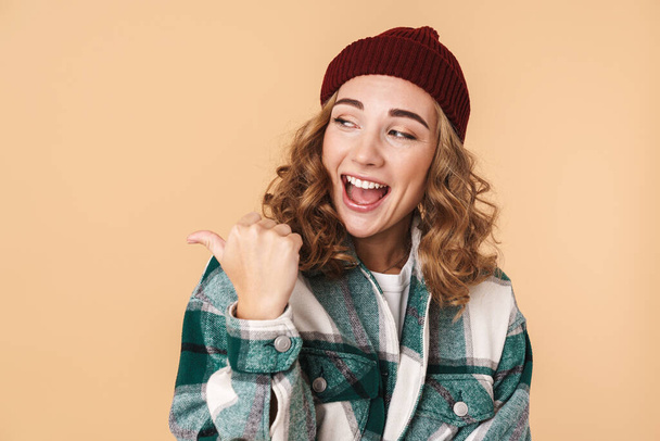 Photo of nice laughing woman in knit hat pointing fingers and looking aside isolated over beige background - Fotó, kép