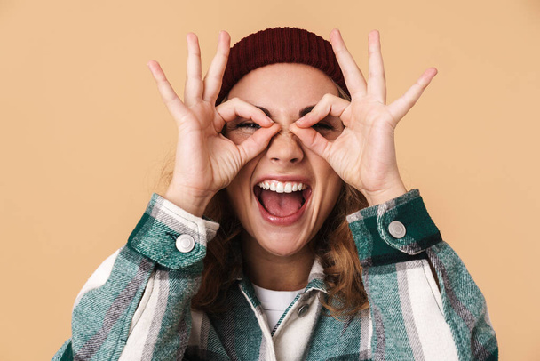 Photo of nice funny woman in knit hat smiling and gesturing ok sign isolated over beige background - Zdjęcie, obraz