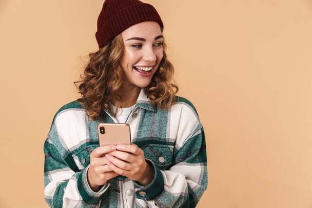 Photo of nice cheerful woman in knit hat smiling and using cellphone isolated over beige background - Zdjęcie, obraz