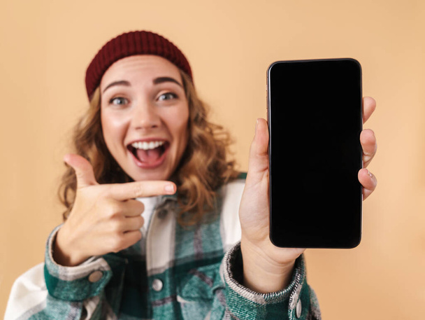 Photo of nice delighted woman in knit hat showing and pointing finger at cellphone isolated over beige background - Fotografie, Obrázek