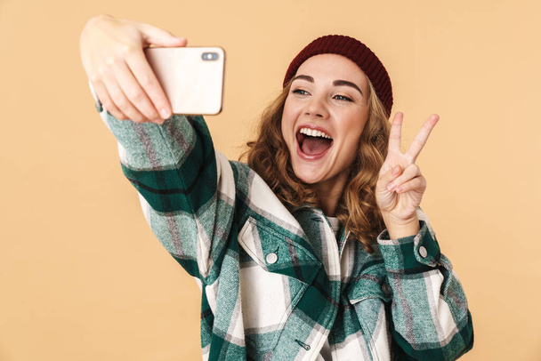 Photo of cheerful woman in knit hat taking selfie on cellphone and gesturing peace sign isolated over beige background - Fotoğraf, Görsel