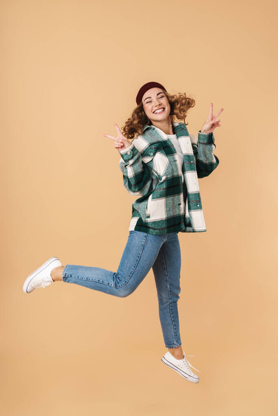 Photo of pretty joyful woman in knit hat jumping and gesturing peace sign isolated over beige background - Φωτογραφία, εικόνα