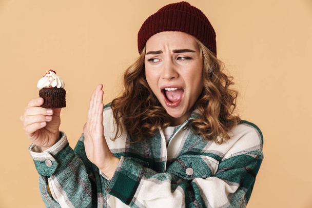 Photo of pretty nervous woman in knit hat making stop gesture and holding cake isolated over beige background - Foto, imagen
