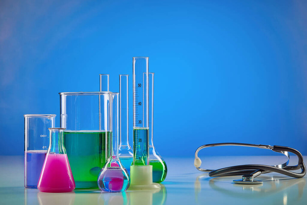Colorful chemical reagents in beakers, medical flasks, measuring cylinder and phonendoscope, blue background. Coronavirus laboratory research. Worldwide pandemic COVID-19. Infectious disease. Close up - Фото, зображення
