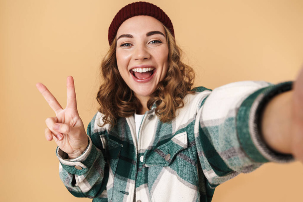 Foto de una mujer bastante feliz en sombrero de punto tomando selfie y haciendo gestos de señal de paz aislado sobre fondo beige
 - Foto, imagen