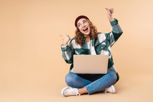 Photo of pretty happy woman pointing fingers aside and using laptop while sitting on floor isolated over beige background - 写真・画像