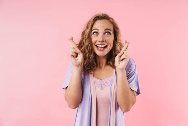 Image of young cheerful woman in stylish pajama smiling and holding fingers crossed isolated over pink background - Foto, Bild