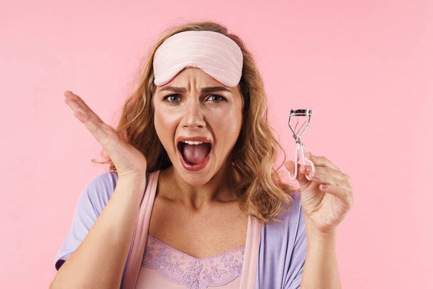 Image of young displeased woman in sleep mask screaming and using eyelashes curler isolated over pink background - Fotografie, Obrázek