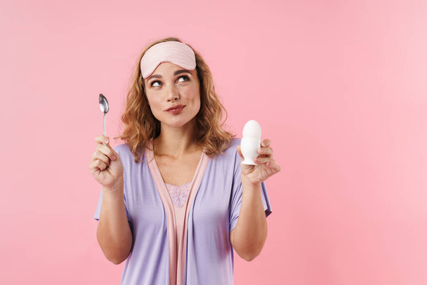 Image of young woman in sleep mask looking upward while holding egg and spoon isolated over pink background - Photo, Image