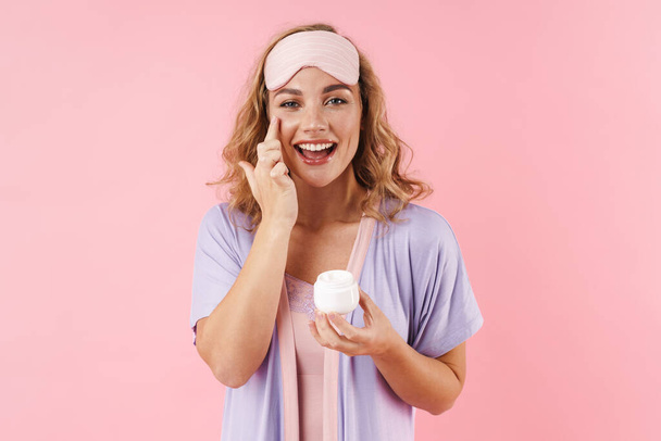 Image of caucasian cheerful woman in sleep mask smiling while applying face cream isolated over pink background - Valokuva, kuva